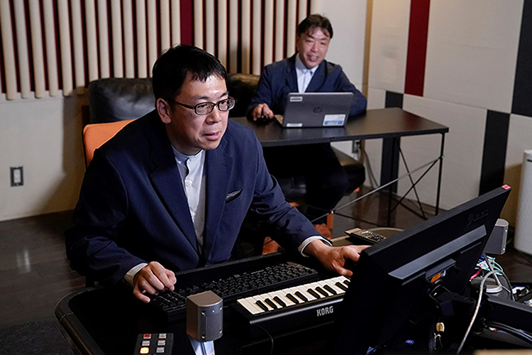 Minamo Takahashi and Hiroyuki Suzuki in suits sitting at desks with computers in a room with blinds.